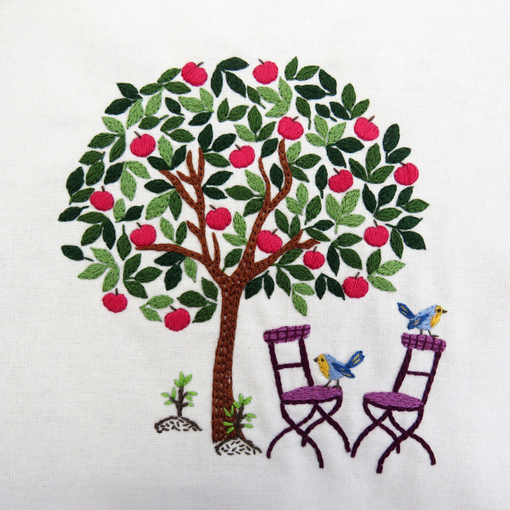 Embroidered design of an apple tree with birds on chairs on a white background