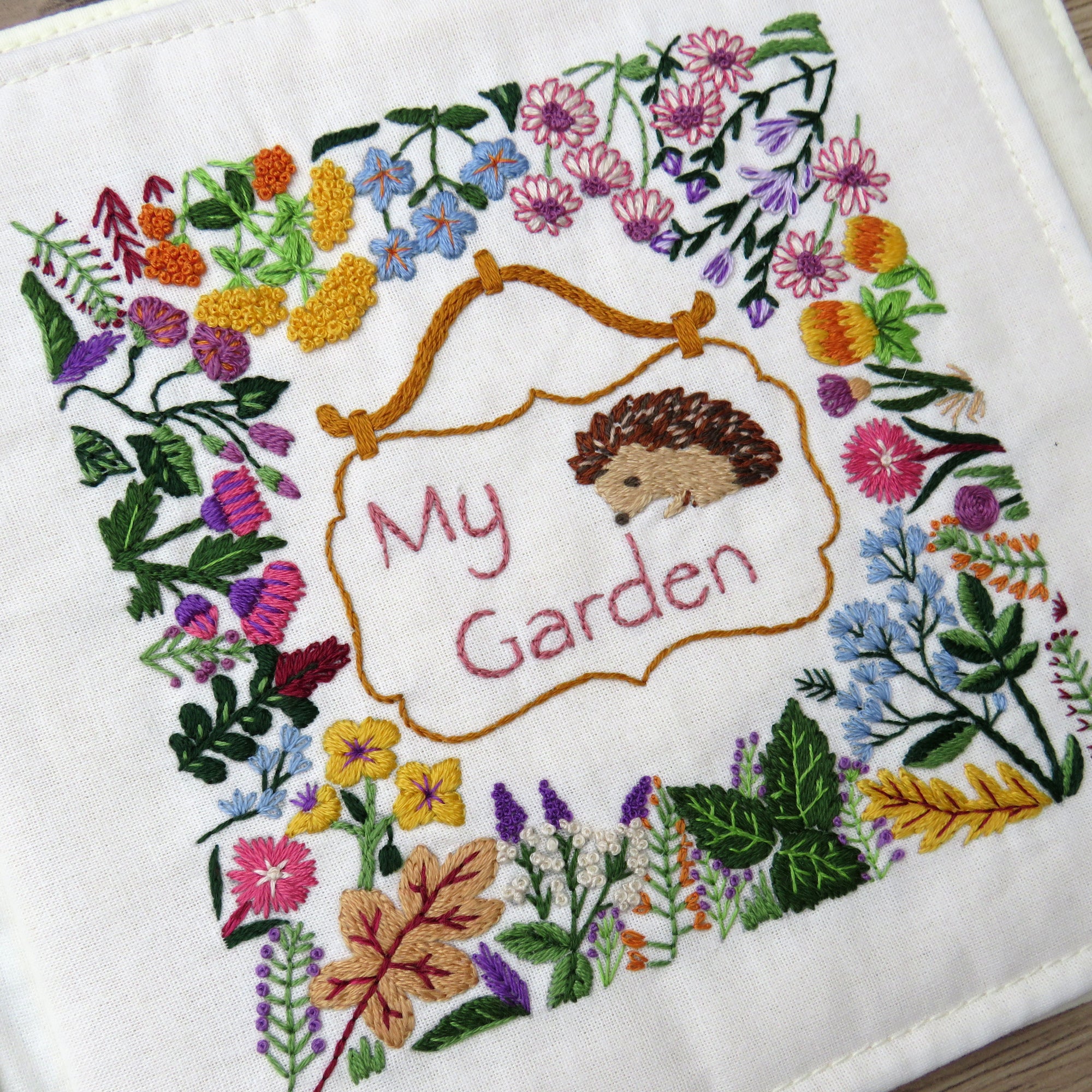 Embroidered design with hedgehog and 'My Garden' text surrounded by colorful flowers and leaves.