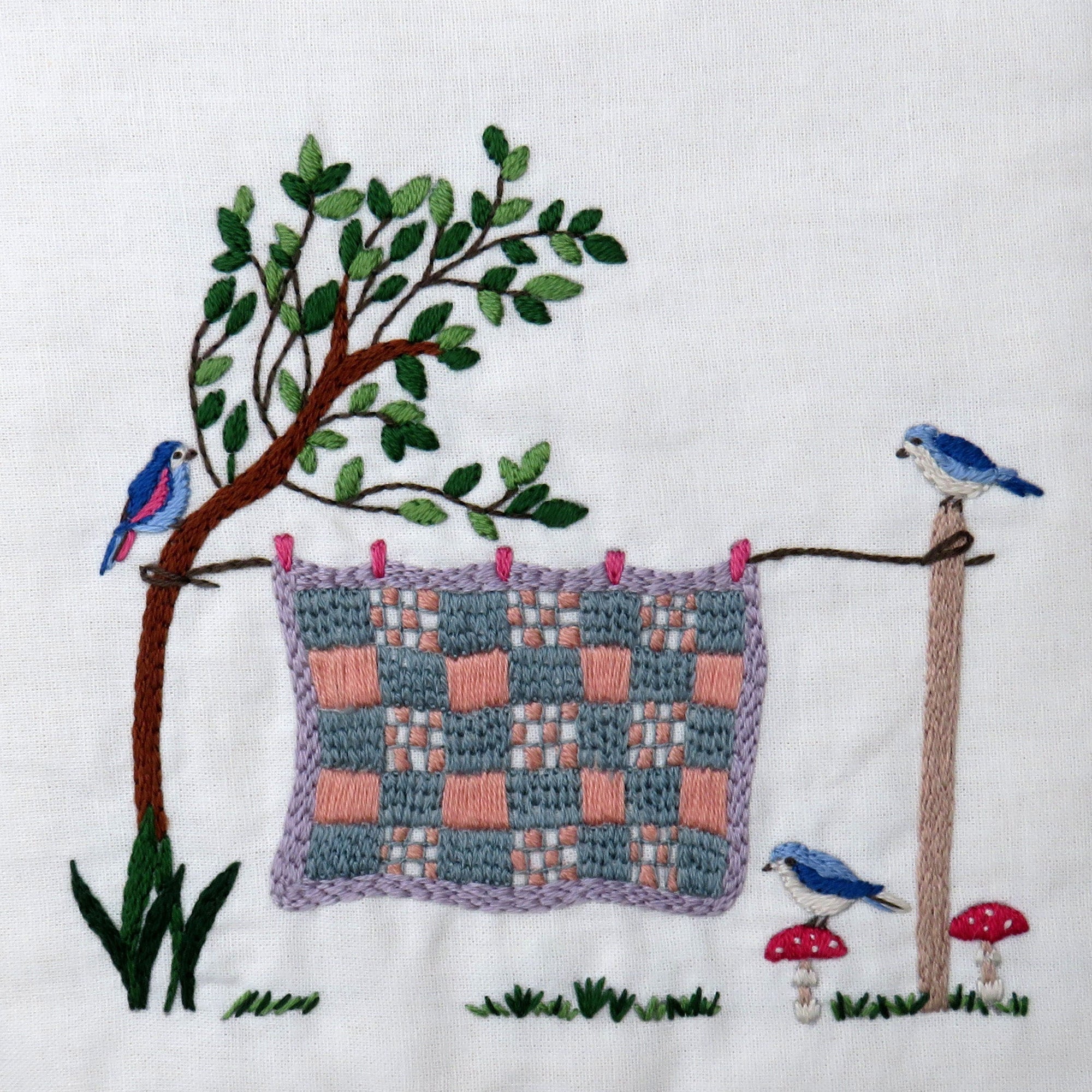 Embroidered scene with a quilt, trees, birds, and mushrooms on a white background.