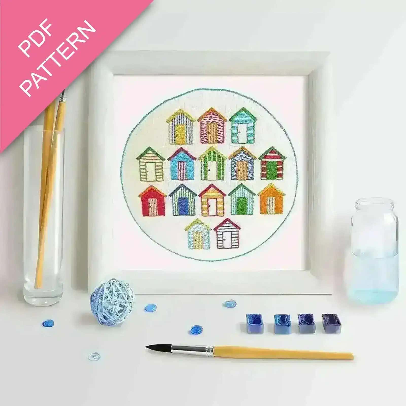 Embroidery kit with colorful beach hut design, thread, and tools on a white background.