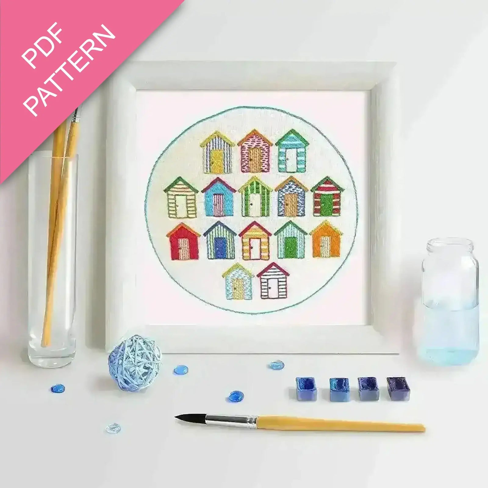 Embroidery kit with colorful beach hut design, thread, and tools on a white background.