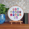 A circular embroidery hoop on a small wooden stand, featuring colorful beach hut embroidery on white fabric. The hoop is displayed on a wooden surface against a gray floral wallpaper background, with a potted green plant on the left and books on the right.