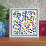 Framed artwork with colorful birds and flowers on a wooden surface with a patterned wall in the background.