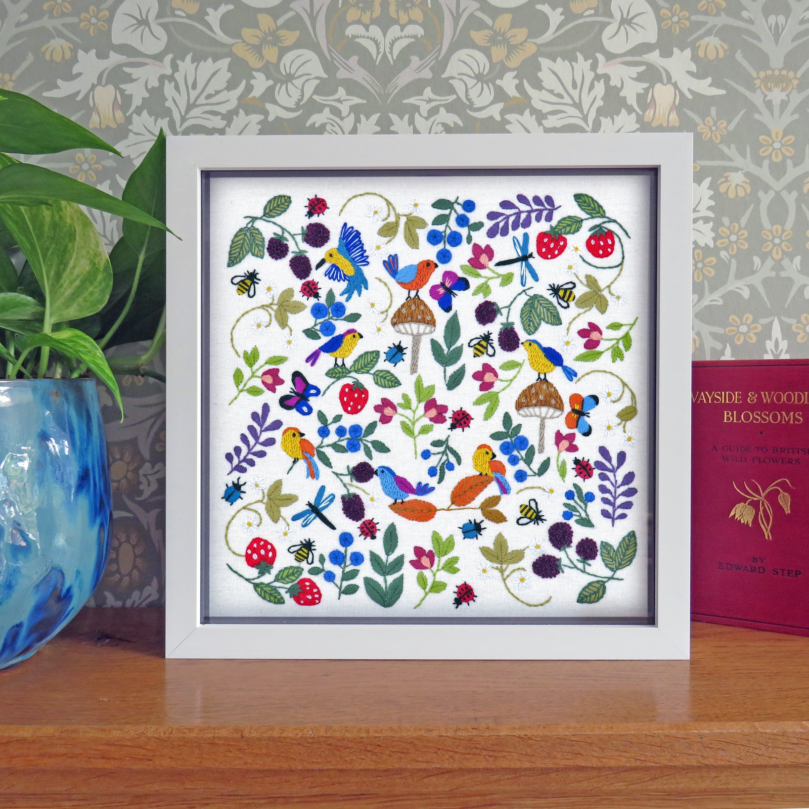 Framed artwork with colorful birds and flowers on a wooden surface with a patterned wall in the background.