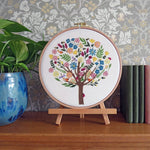 Embroidery hoop with a colorful floral tree design on a wooden stand against a floral wallpapered wall.