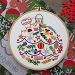 A round embroidery hoop holding a white fabric panel with colorful Christmas-themed embroidery, including ornaments, stars, candy canes, snowflakes, and a 'Merry Christmas' tag, displayed on a patterned holiday textile.