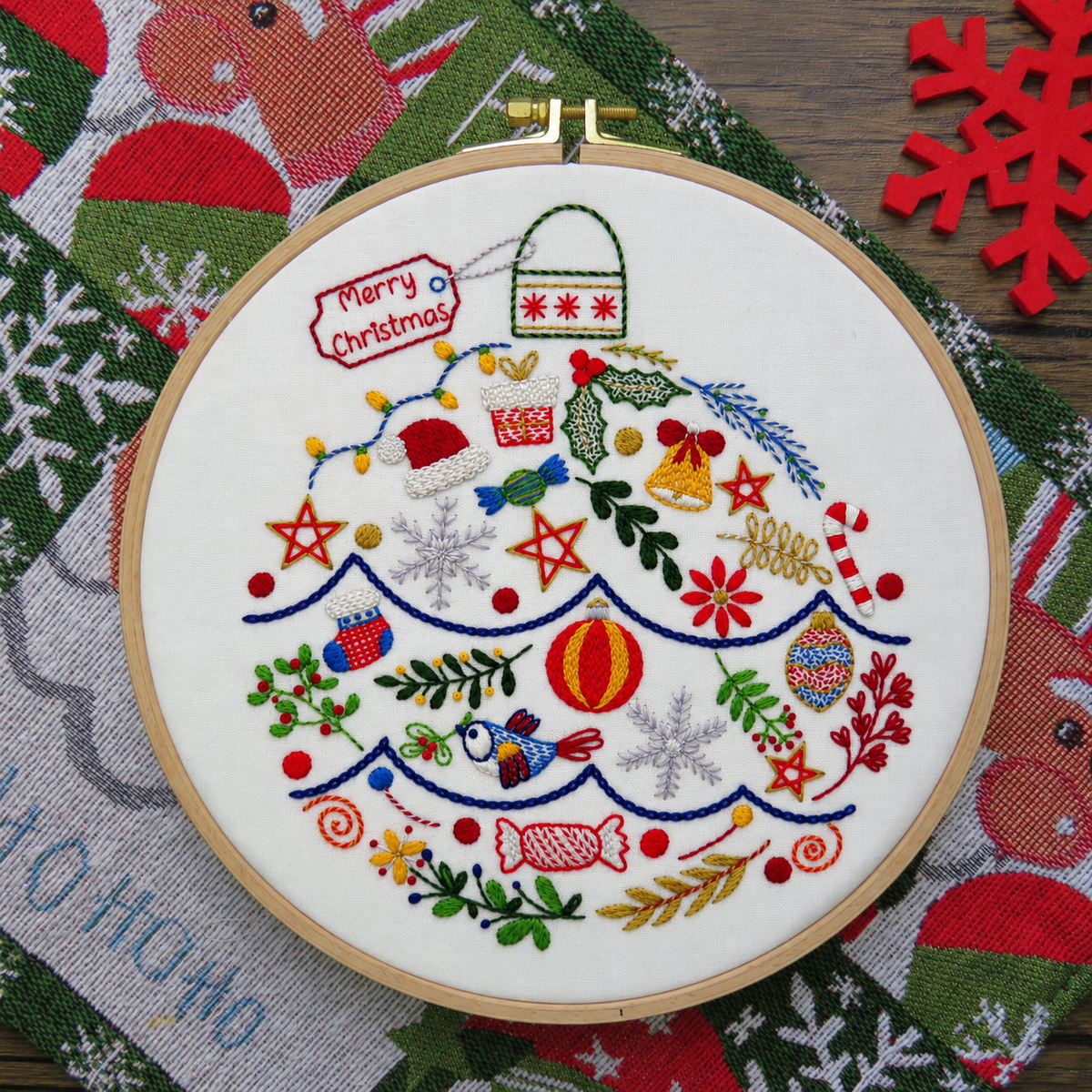 A round embroidery hoop holding a white fabric panel with colorful Christmas-themed embroidery, including ornaments, stars, candy canes, snowflakes, and a 'Merry Christmas' tag, displayed on a patterned holiday textile.