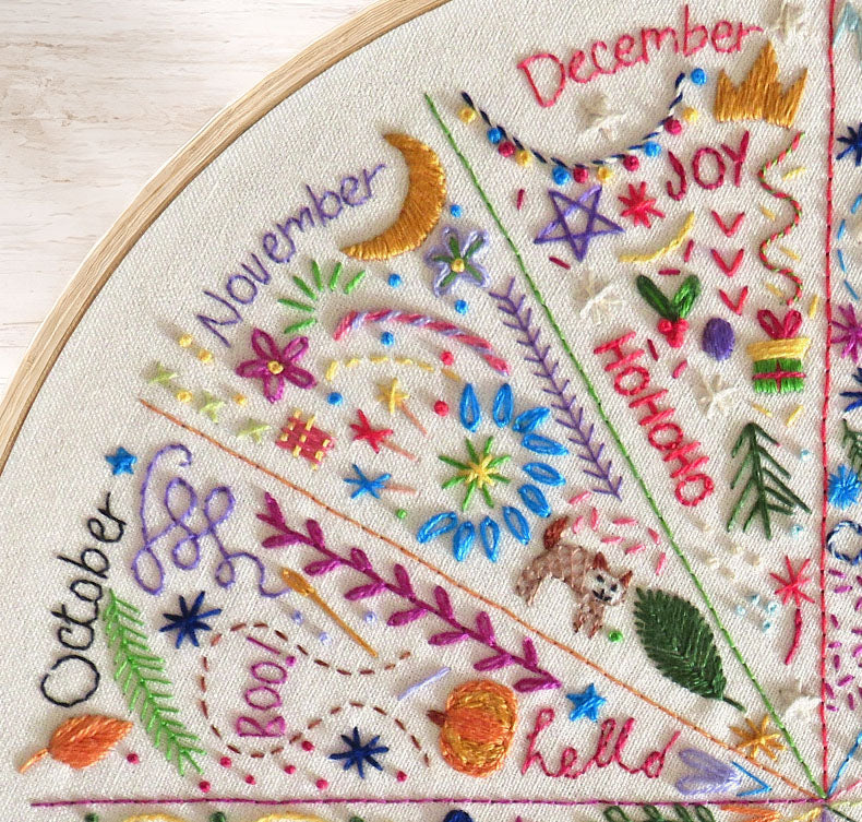 A close-up of a circular embroidery hoop containing a detailed seasonal floral and celestial embroidery design. The design features embroidered text for 'October', 'November', 'December', and 'Hello', along with stars, moons, leaves, flowers, and small animals like a rabbit and squirrel stitched on a cream fabric background.