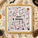 A framed cross-stitch design featuring a colorful 'Happy Halloween' pattern with pumpkins, bats, spiderwebs, ghosts, and witch motifs, styled on a patterned fabric background with seasonal props.