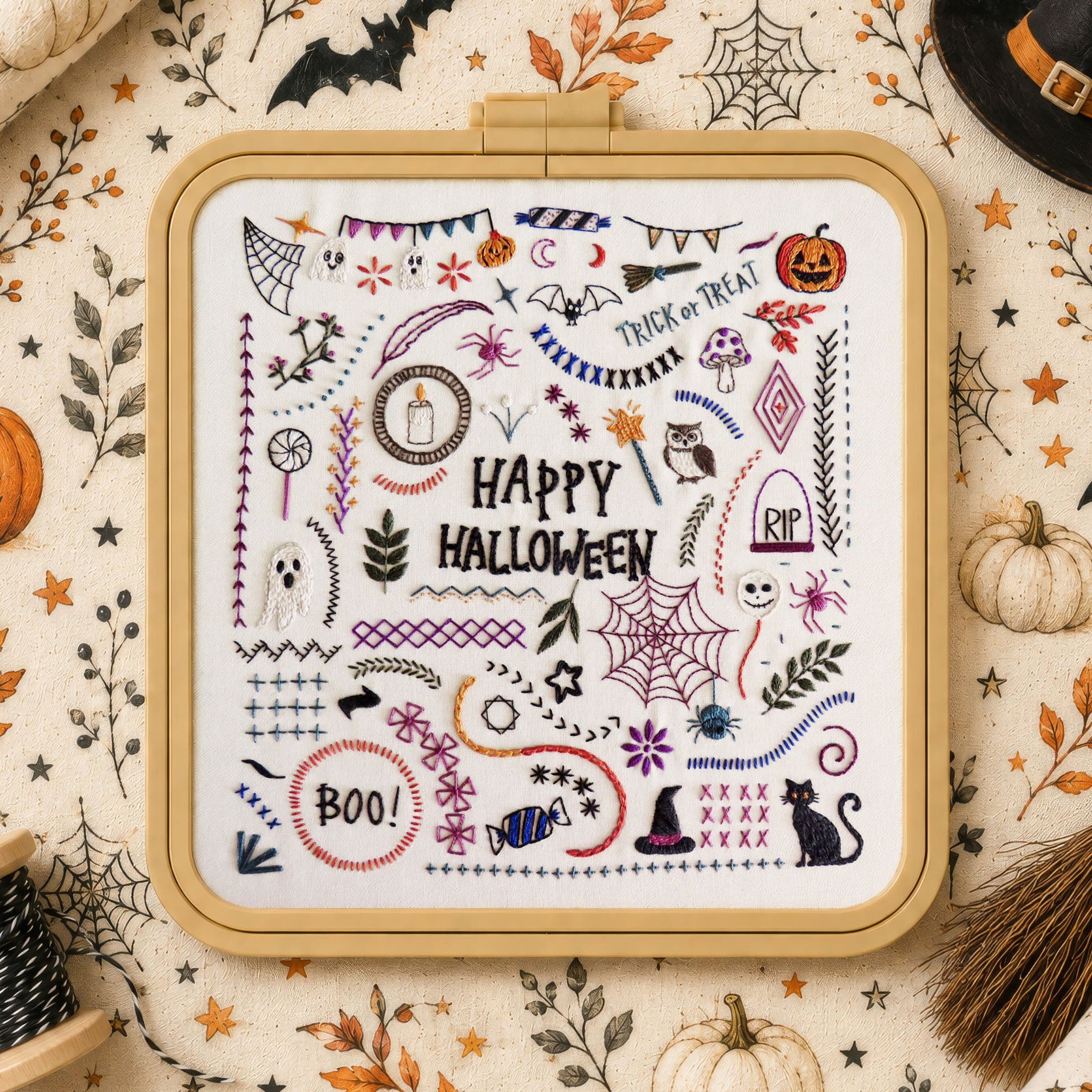A framed cross-stitch design featuring a colorful 'Happy Halloween' pattern with pumpkins, bats, spiderwebs, ghosts, and witch motifs, styled on a patterned fabric background with seasonal props.