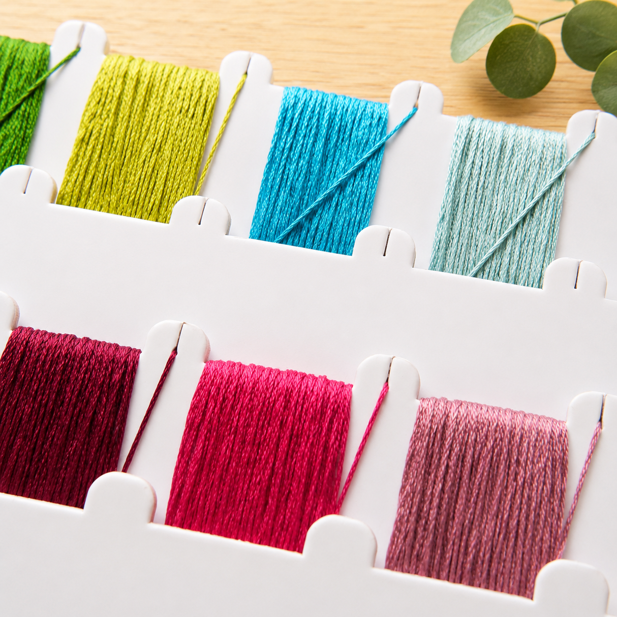A white embroidery thread spool organizer holding multiple colorful threads in green, blue, light blue, burgundy, pink, and mauve, arranged on a light wood surface with a small green leaf sprig in the corner.