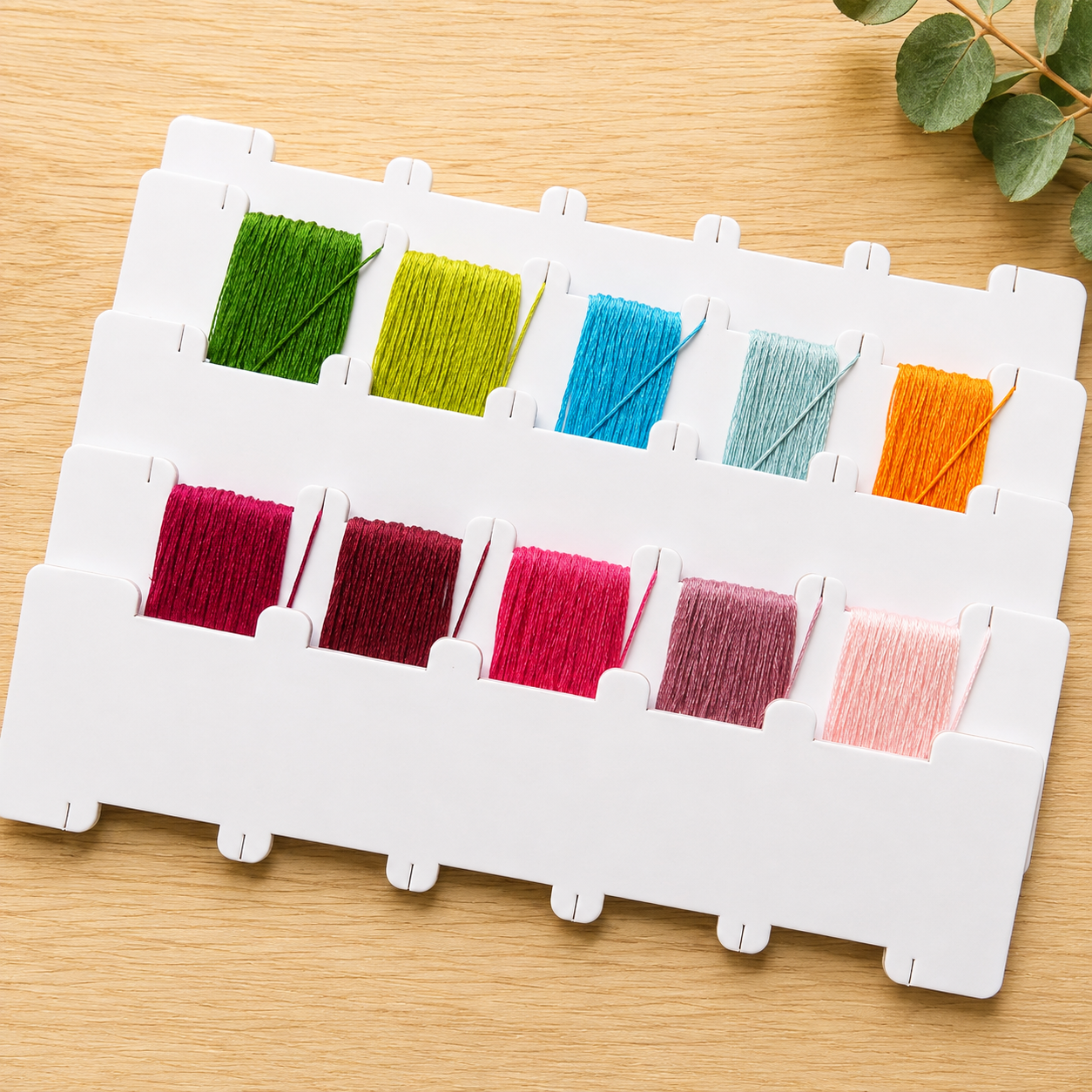 A white plastic embroidery floss display board holding eight spools of thread in assorted colors, arranged on a light wood surface with a small green leafy sprig in the corner.