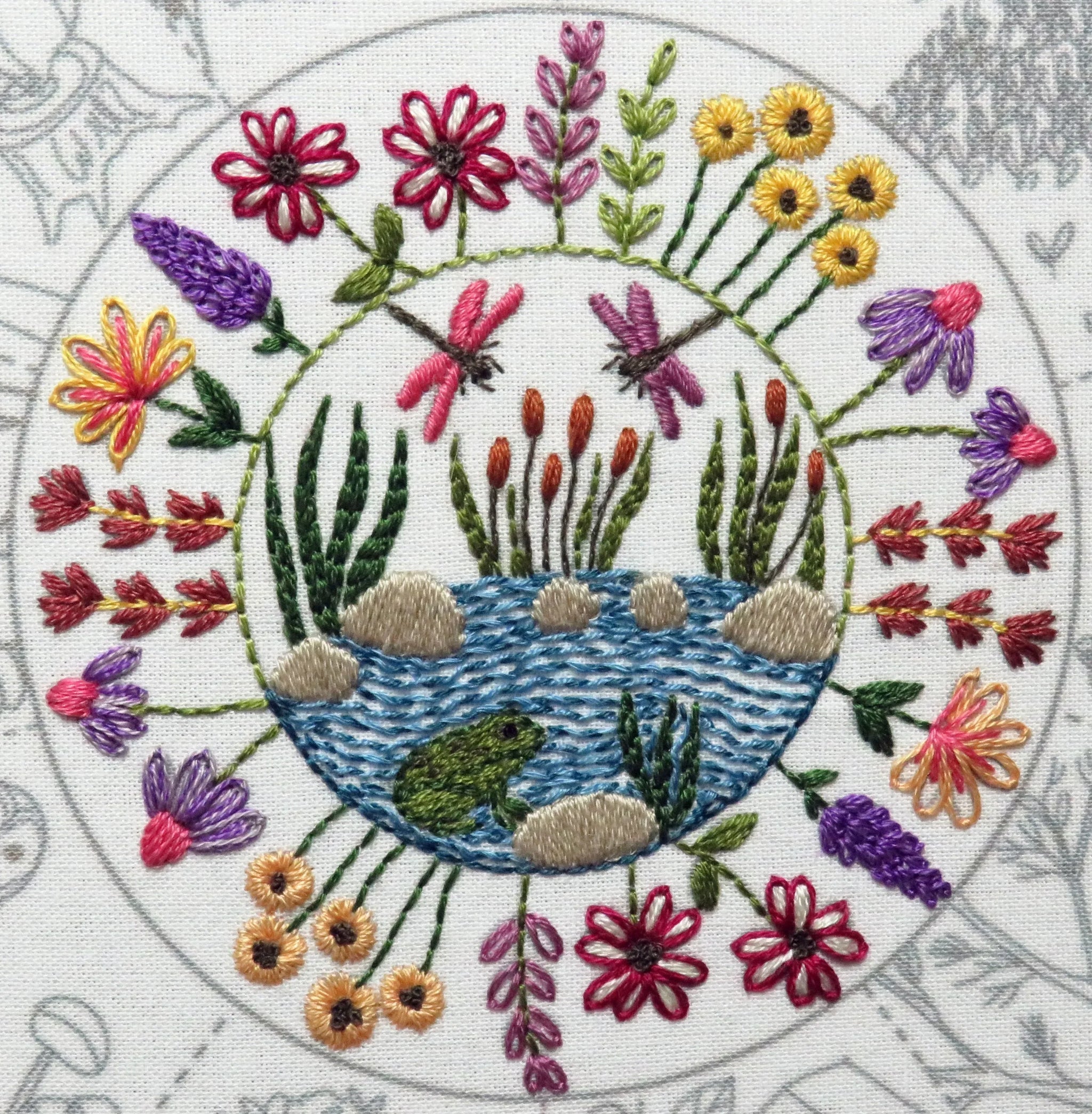 Embroidered design of a pond with flowers and dragonflies on a circular pattern.