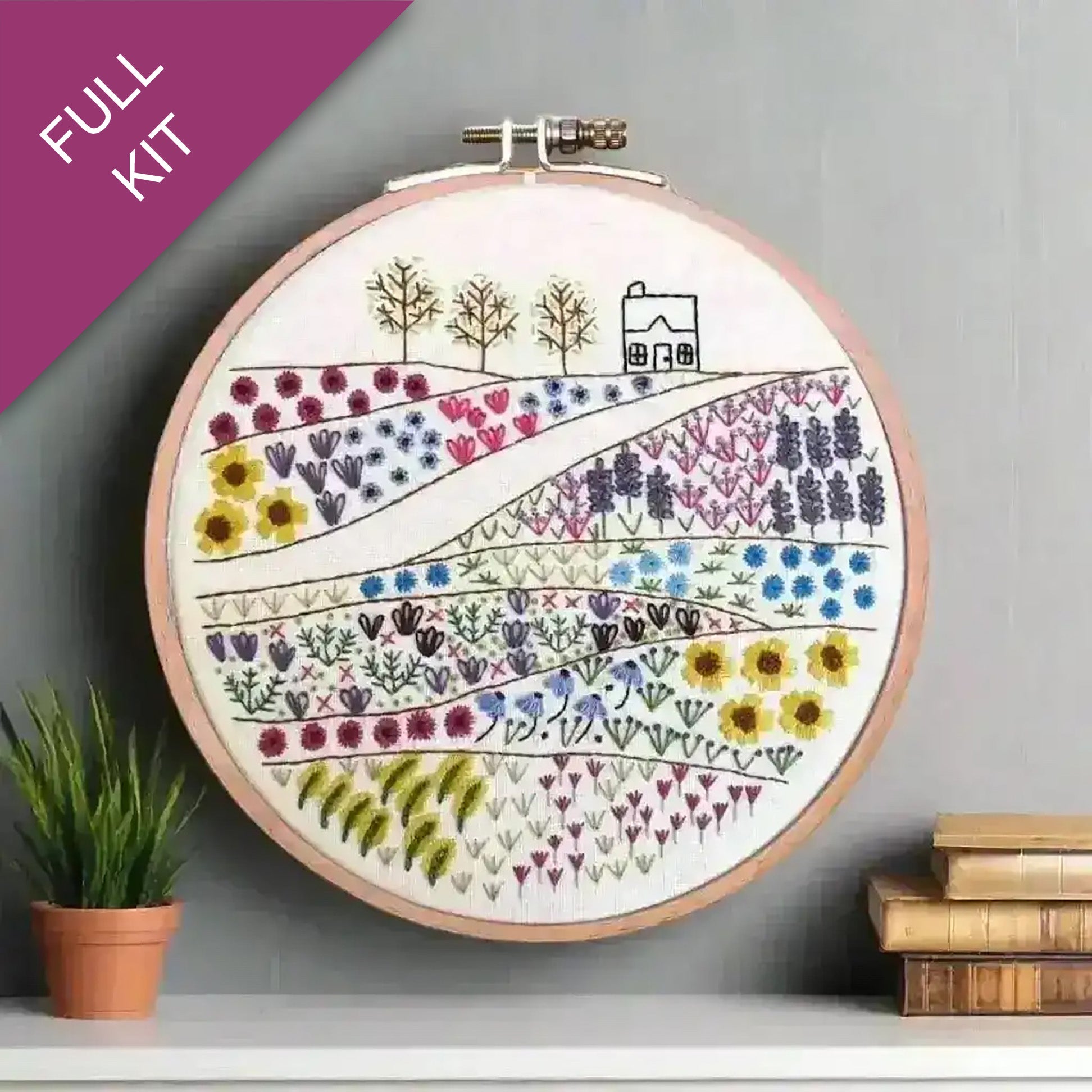 Embroidery hoop with a colorful landscape design on a gray surface with a plant and books.
