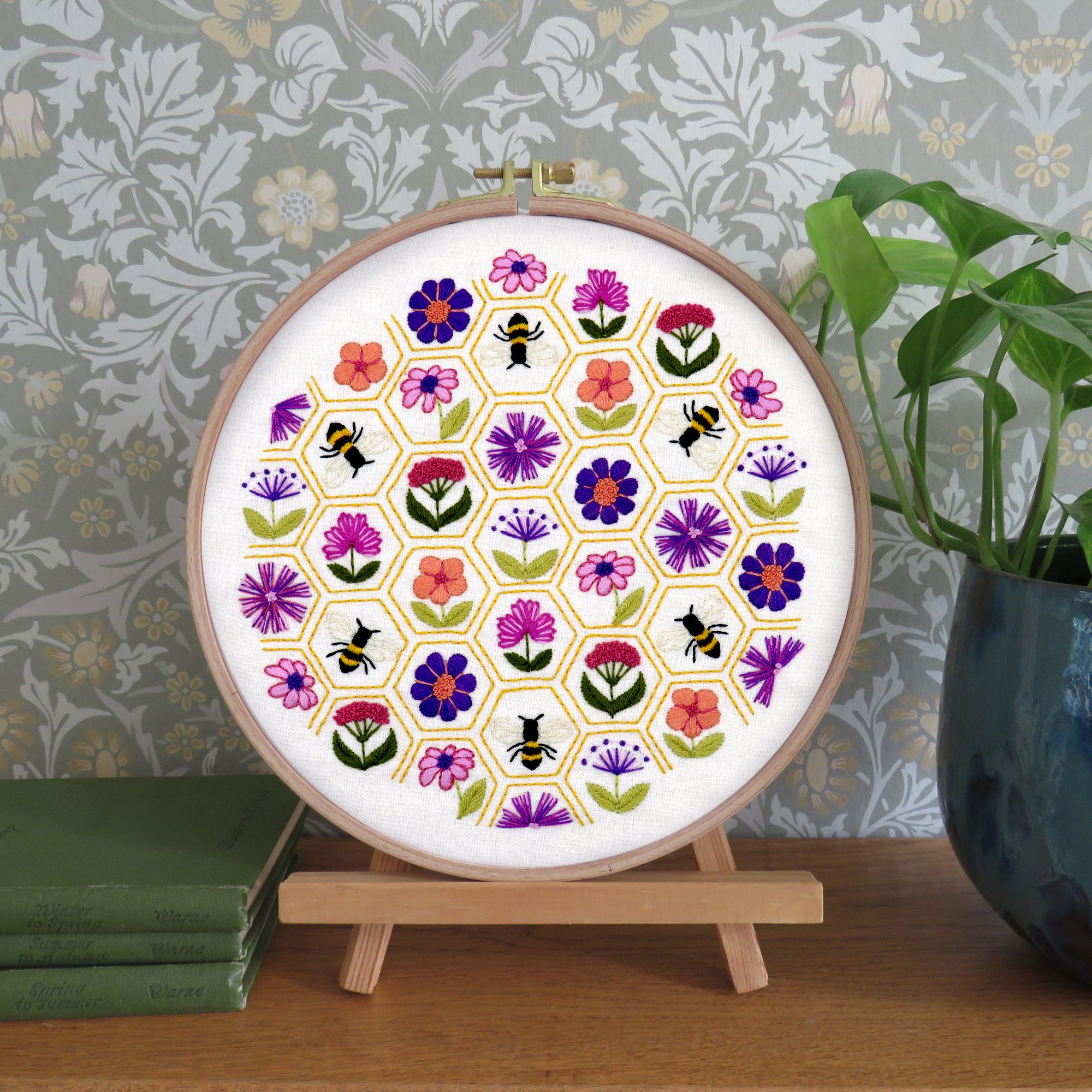 A circular embroidery hoop displaying a colorful floral and bee pattern, mounted on a small wooden stand on a tabletop. The background features a gray floral wallpaper, a green plant in a blue pot, and a stack of green books.