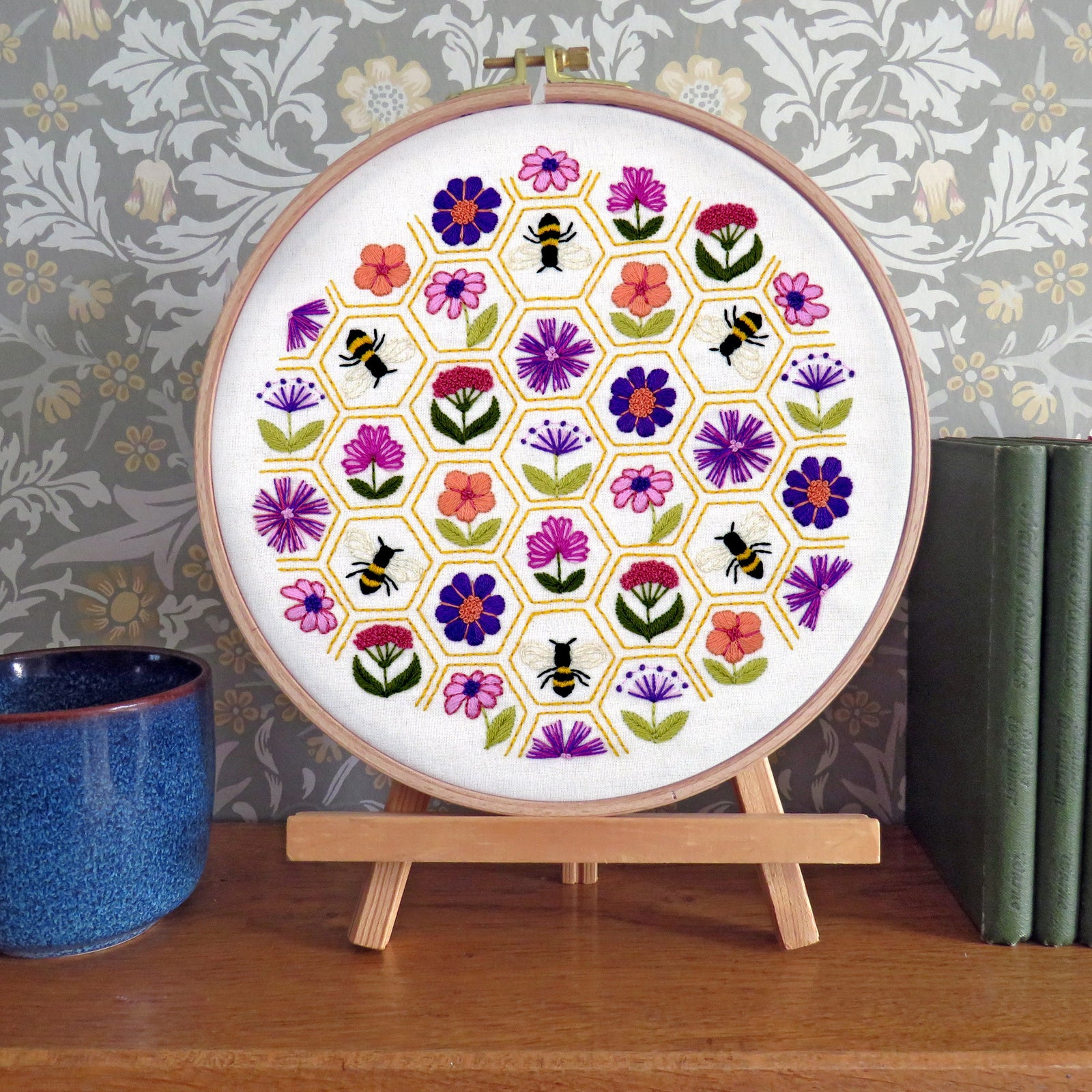 Embroidery hoop with floral and bee pattern on a wooden stand against a decorative wall.
