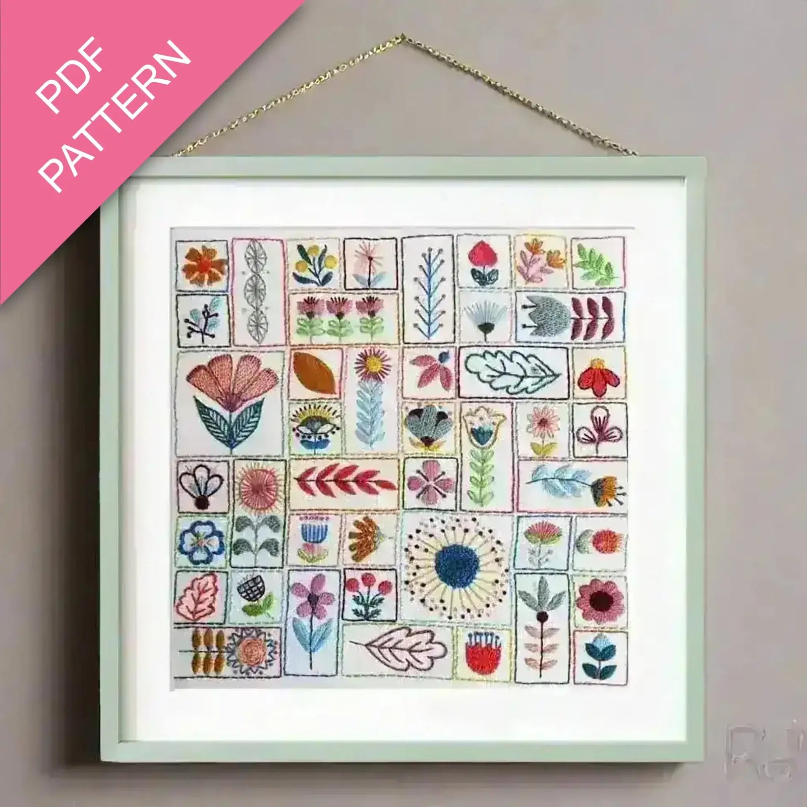 Framed embroidery pattern with floral designs on a plain wall, featuring a pink PDF pattern overlay.