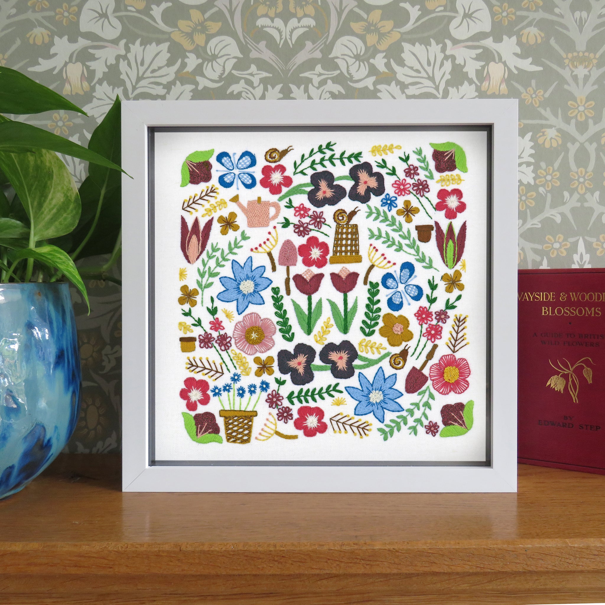 A framed floral embroidery or needlework artwork displayed on a wooden surface, with a potted plant on the left and a red book on the right. The artwork features a dense arrangement of colorful flowers, leaves, and small decorative motifs inside a white frame.