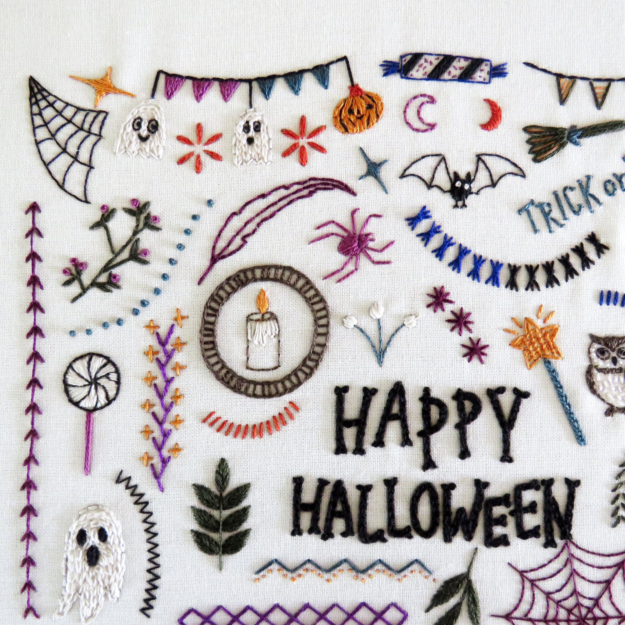 A close-up of a white fabric featuring colorful embroidered Halloween motifs and the words “HAPPY HALLOWEEN” stitched in black.