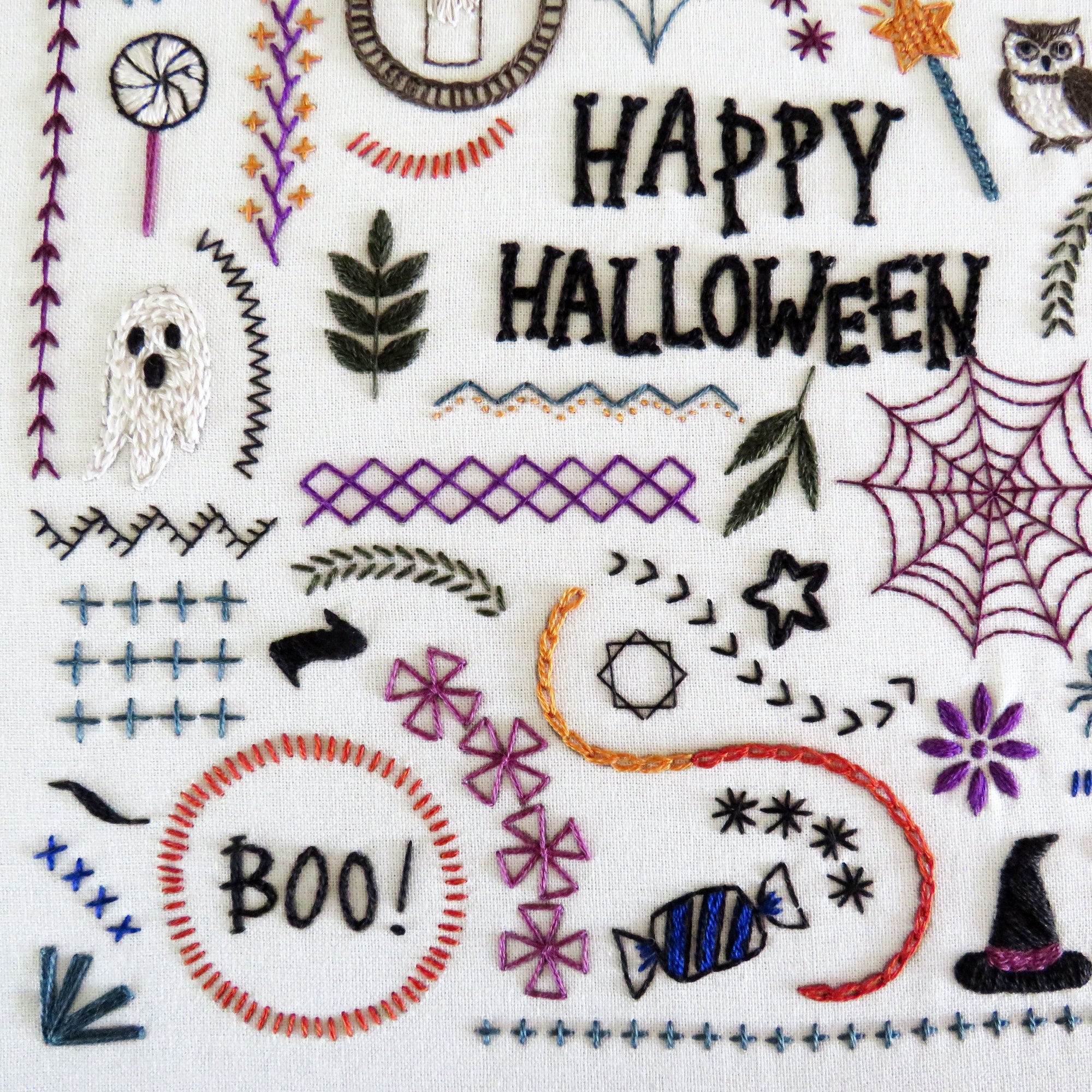 A close-up of a white fabric featuring colorful embroidered Halloween-themed designs, including the words 'HAPPY HALLOWEEN' and 'BOO!', a ghost, spiderweb, candy, witch hat, leaves, stars, and decorative stitch patterns.