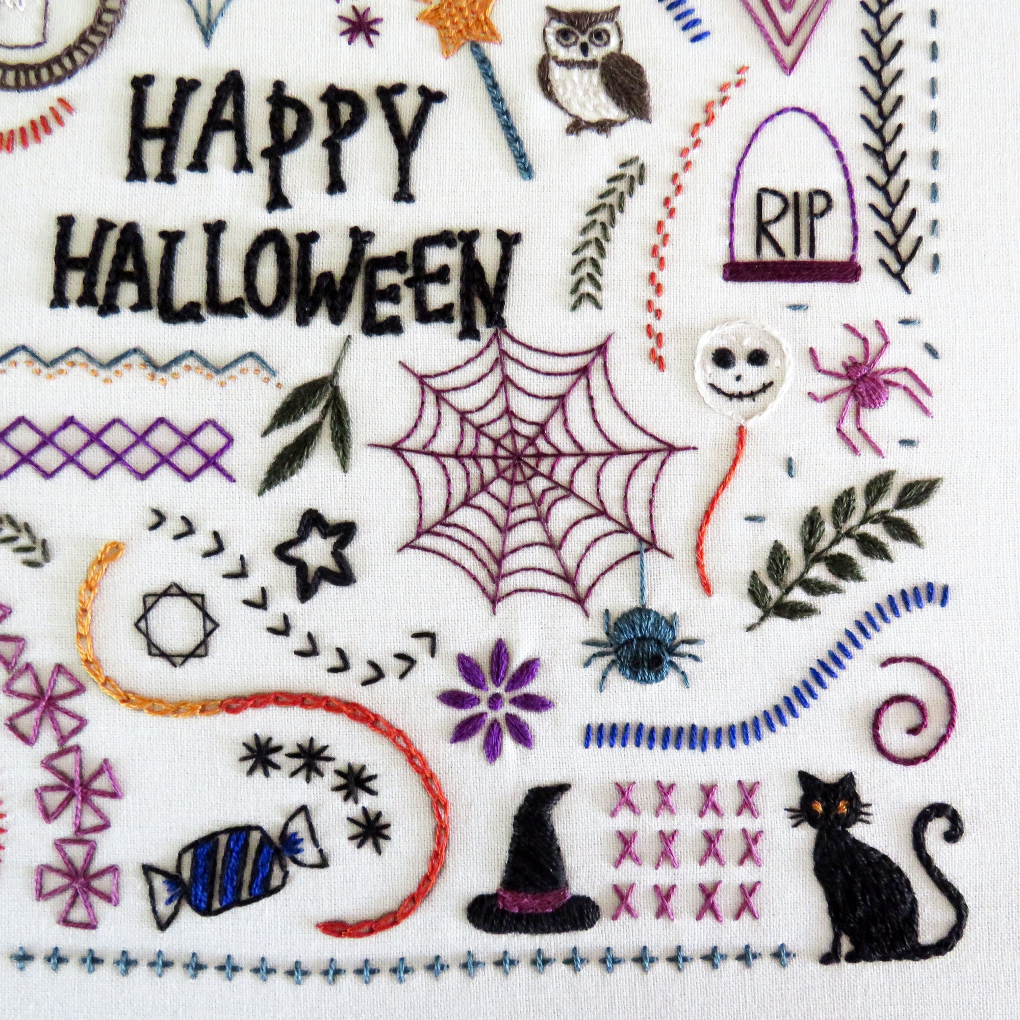 A close-up of a white fabric featuring colorful embroidered Halloween-themed designs, including the words 'HAPPY HALLOWEEN,' a spiderweb, spider, witch hat, black cat, candy, leaves, stars, and decorative borders.