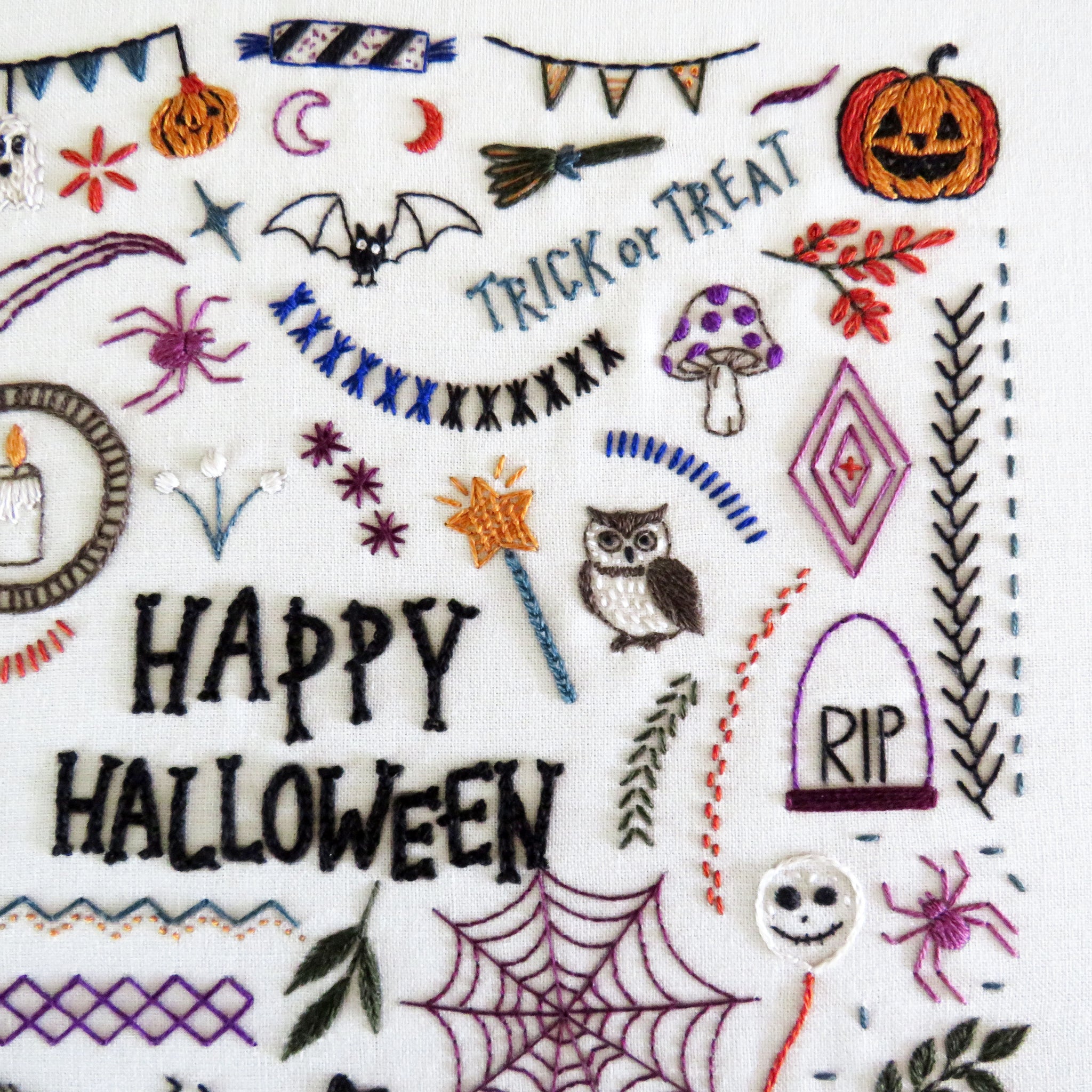 Close-up of a white fabric surface with embroidered Halloween motifs and the words “HAPPY HALLOWEEN” in black thread, including a jack-o'-lantern, spider, moon, broom, witch hat, spiderweb, owl, mushrooms, stars, and decorative borders.
