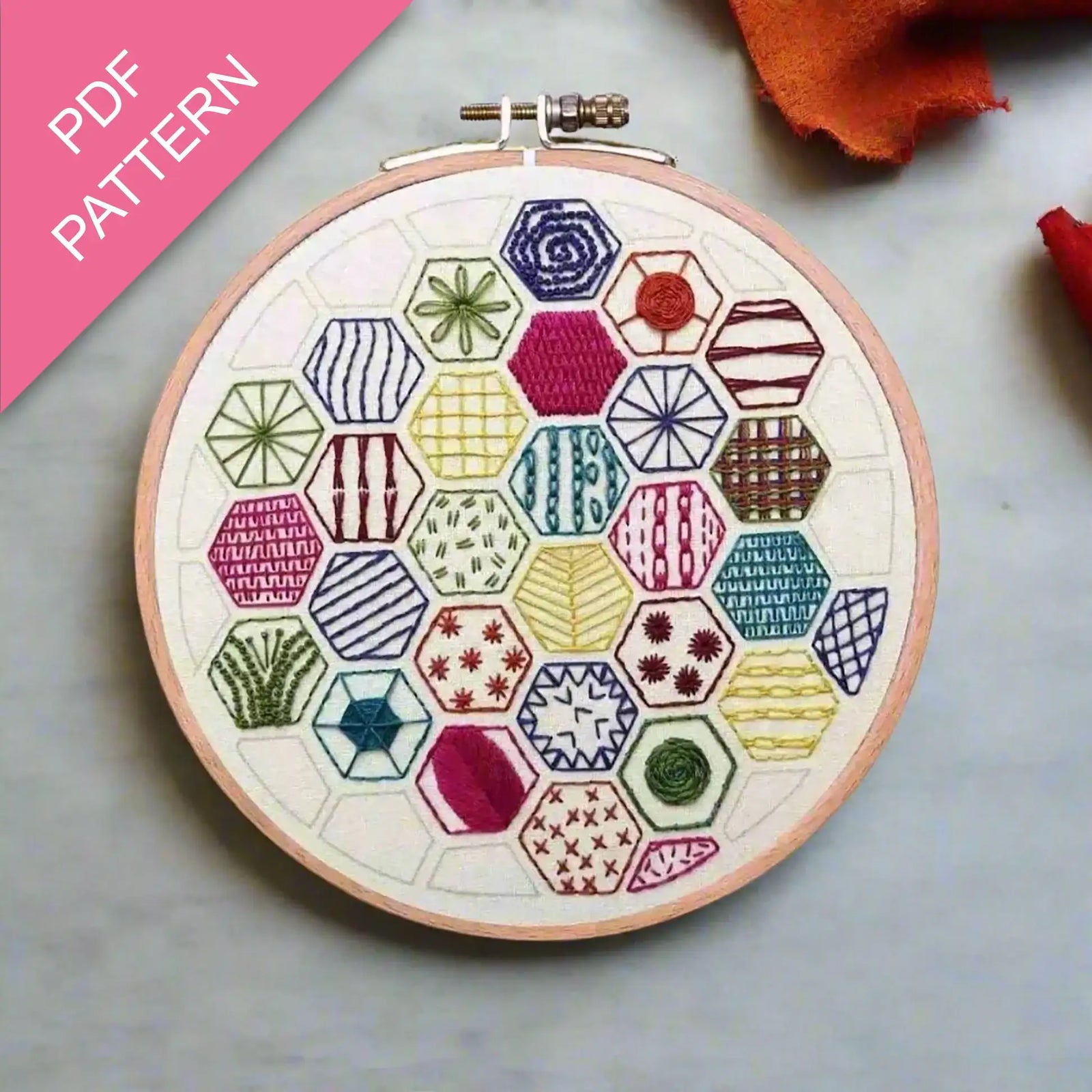 Embroidery hoop with colorful geometric pattern on a gray surface, with 'PDF Pattern' text on a pink background.