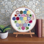 A circular embroidery hoop displaying a colorful geometric hexagon pattern, mounted on a small wooden stand on a wooden table. The background features floral wallpaper, a small potted plant on the left, and vintage books on the right.