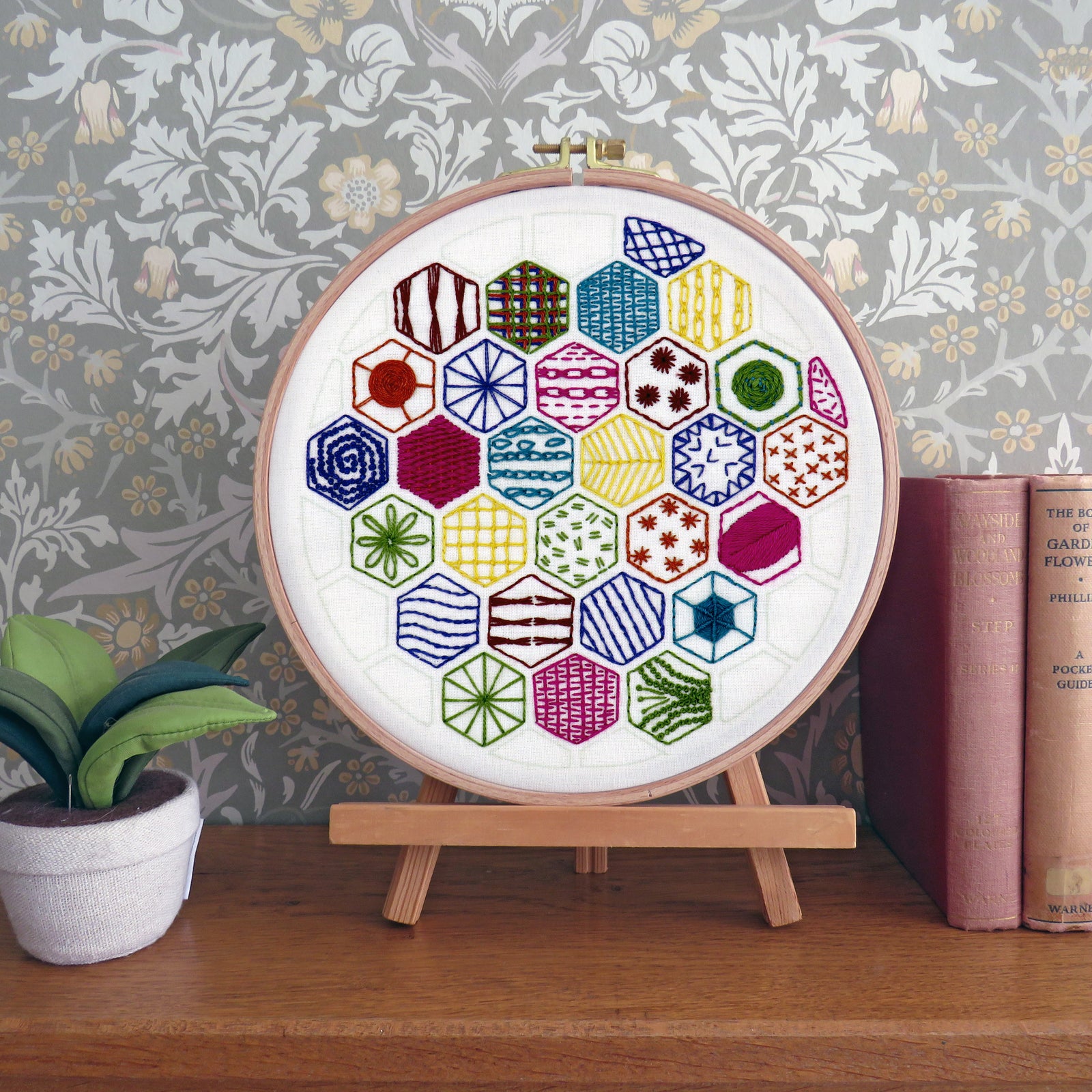 A circular embroidery hoop displaying a colorful geometric hexagon pattern, mounted on a small wooden stand on a wooden table. The background features floral wallpaper, a small potted plant on the left, and vintage books on the right.