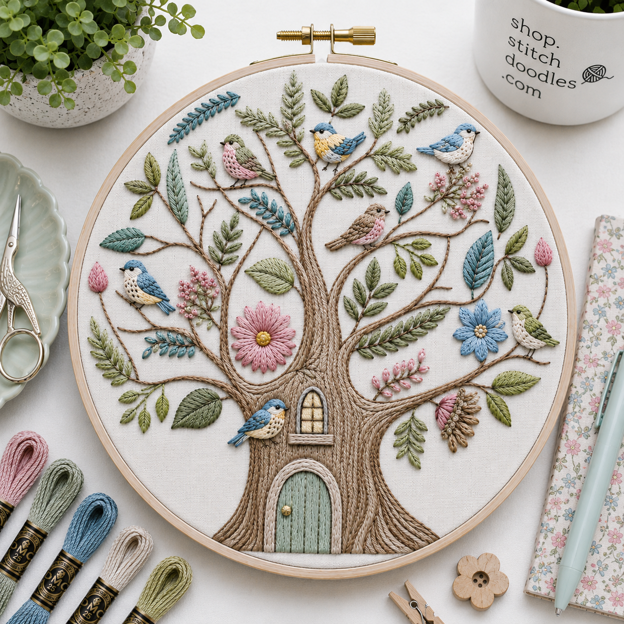 Embroidery hoop with a tree design featuring birds and flowers, surrounded by embroidery supplies on a white surface.