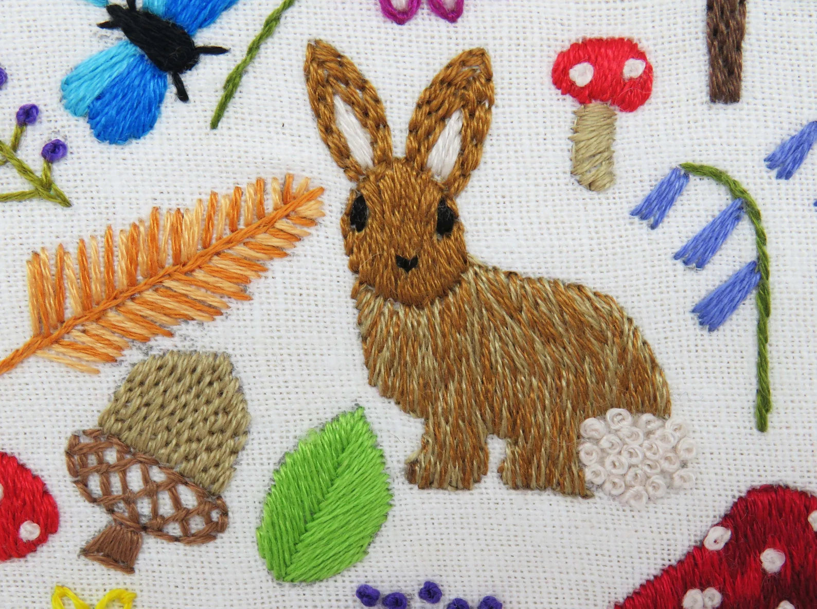 Embroidered design of a rabbit surrounded by flowers, leaves, and mushrooms on a white background.