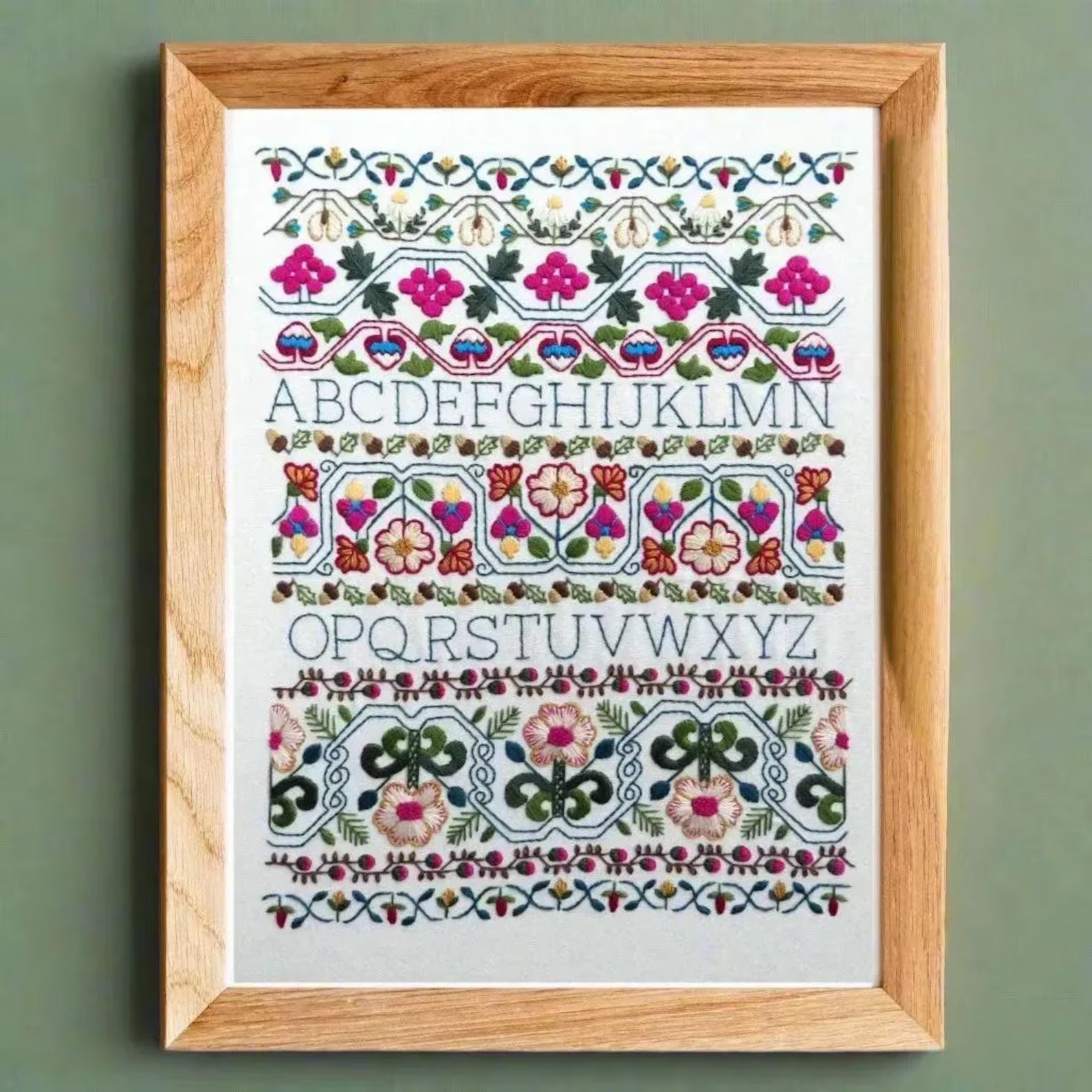 Framed embroidery with floral and geometric patterns and alphabet letters on a green wall.