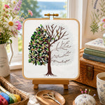 Cross-stitching project with a tree design on a wooden table with flowers and books in the background.