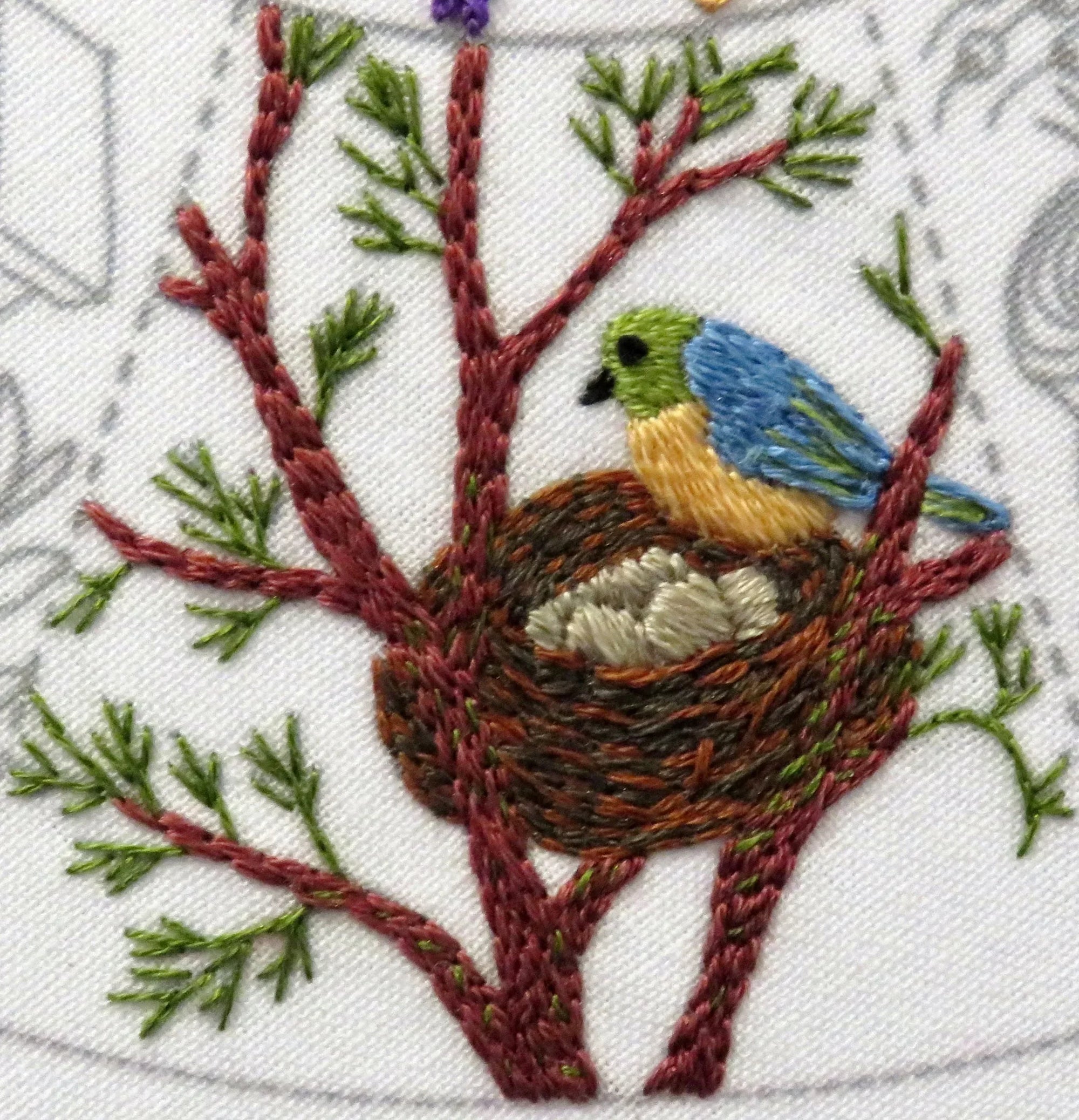 Embroidered design of a bird in a nest on a branch with green leaves.