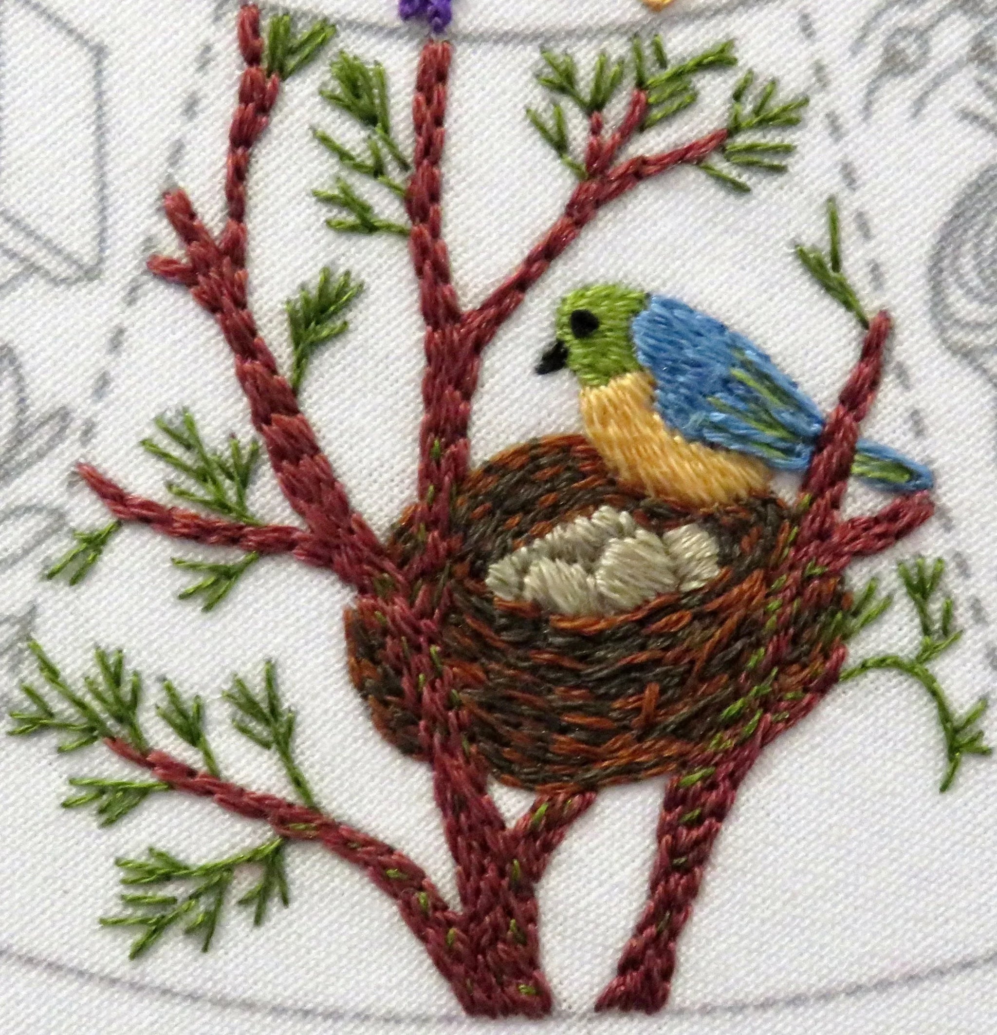 Embroidered design of a bird in a nest on a branch with green leaves.