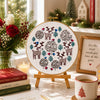 Decorative embroidery hoop with Christmas-themed design on a table with books, flowers, and a mug.