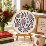 Decorative embroidery hoop with Christmas-themed design on a table with books, flowers, and a mug.
