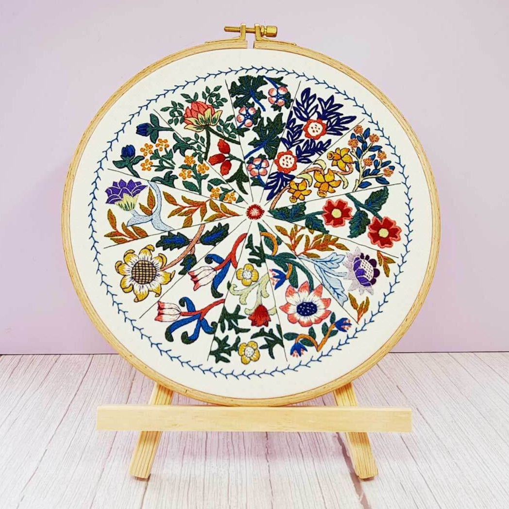 Embroidery hoop with colorful floral pattern on a light wooden stand against a light pink background