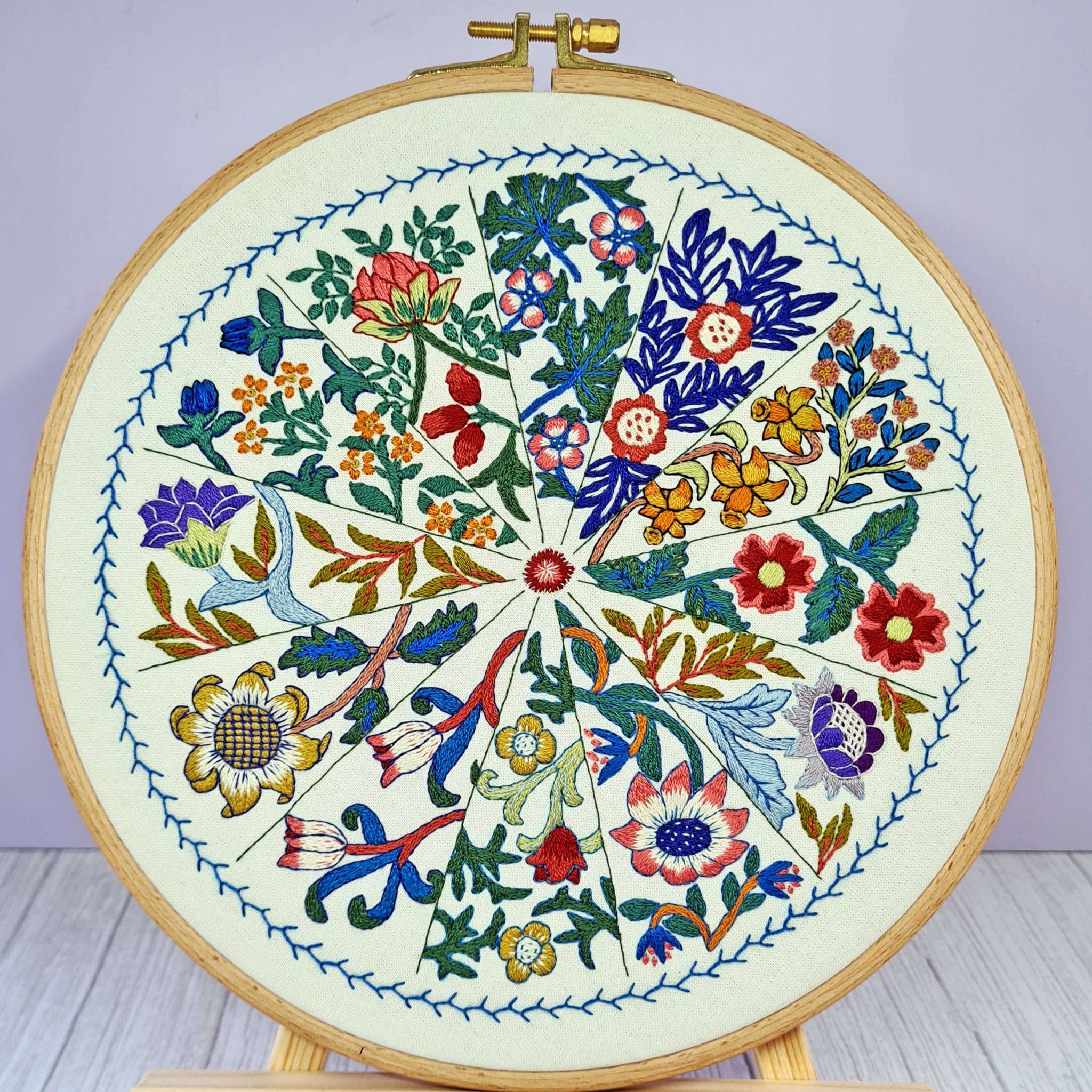 Embroidery hoop with colorful floral design on a wooden stand against a light purple background