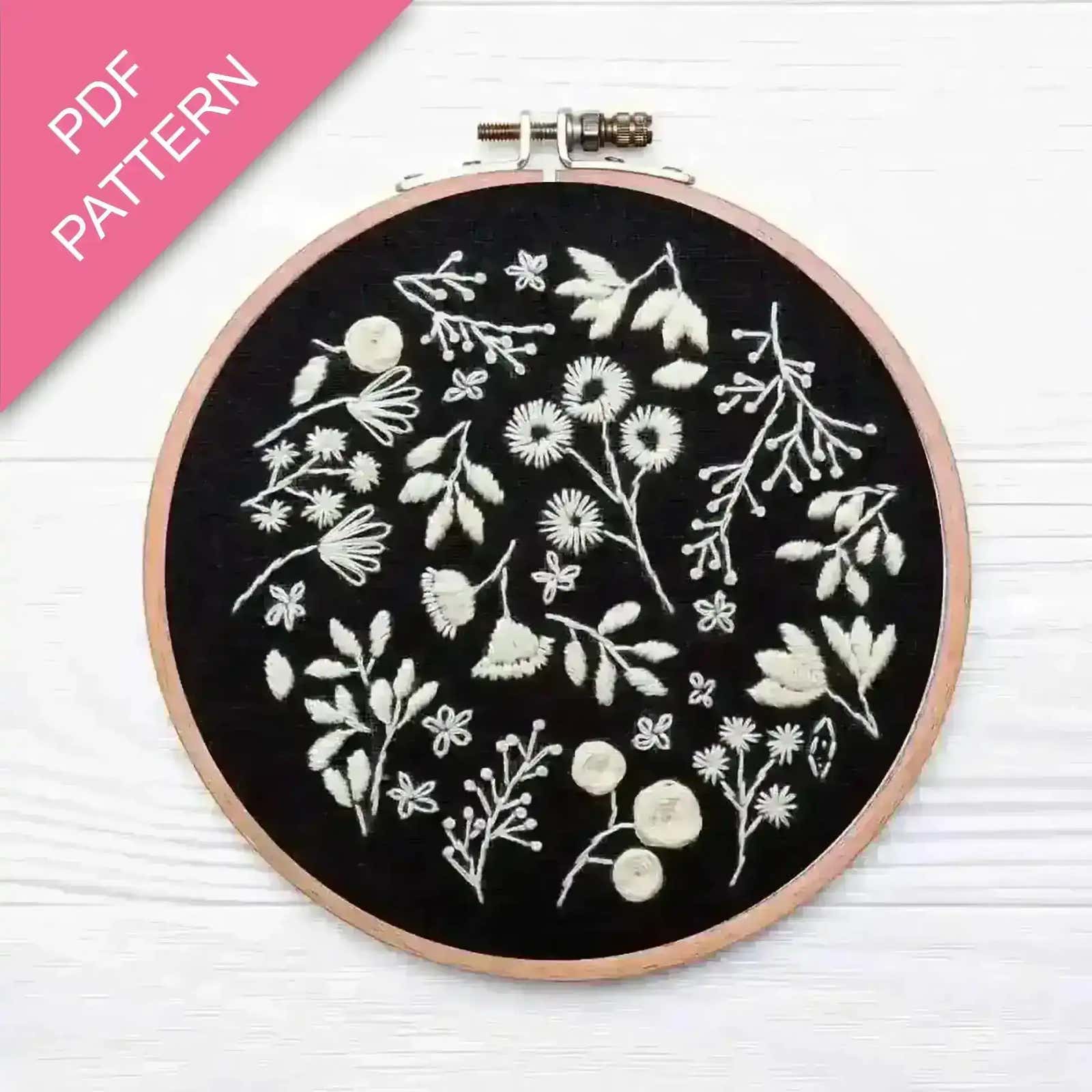 Embroidery hoop with white floral patterns on black fabric, pink PDF pattern label.