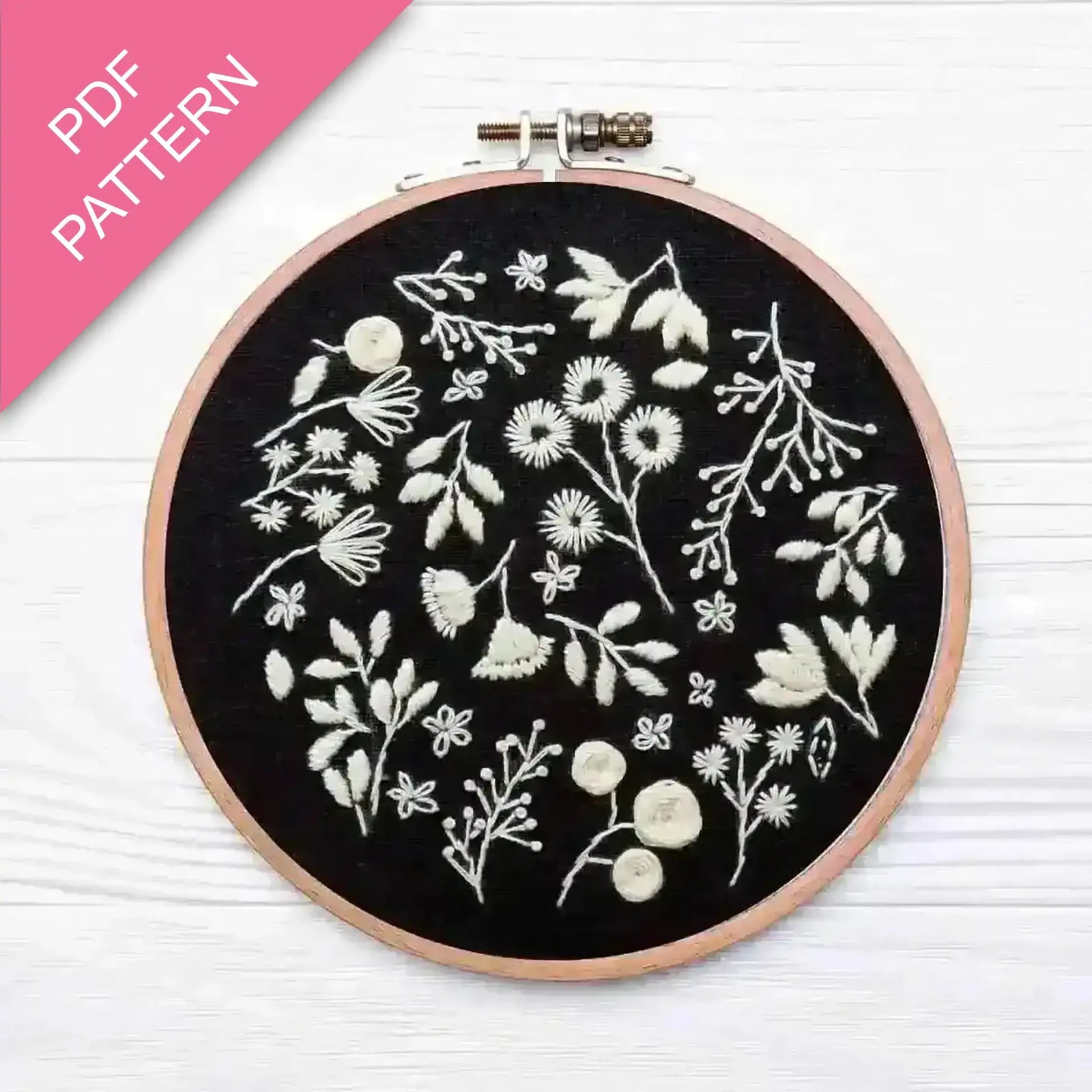 Embroidery hoop with white floral patterns on black fabric, pink PDF pattern label.