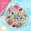 Embroidery hoop with colorful underwater scene including fish and coral on a blue wooden surface.