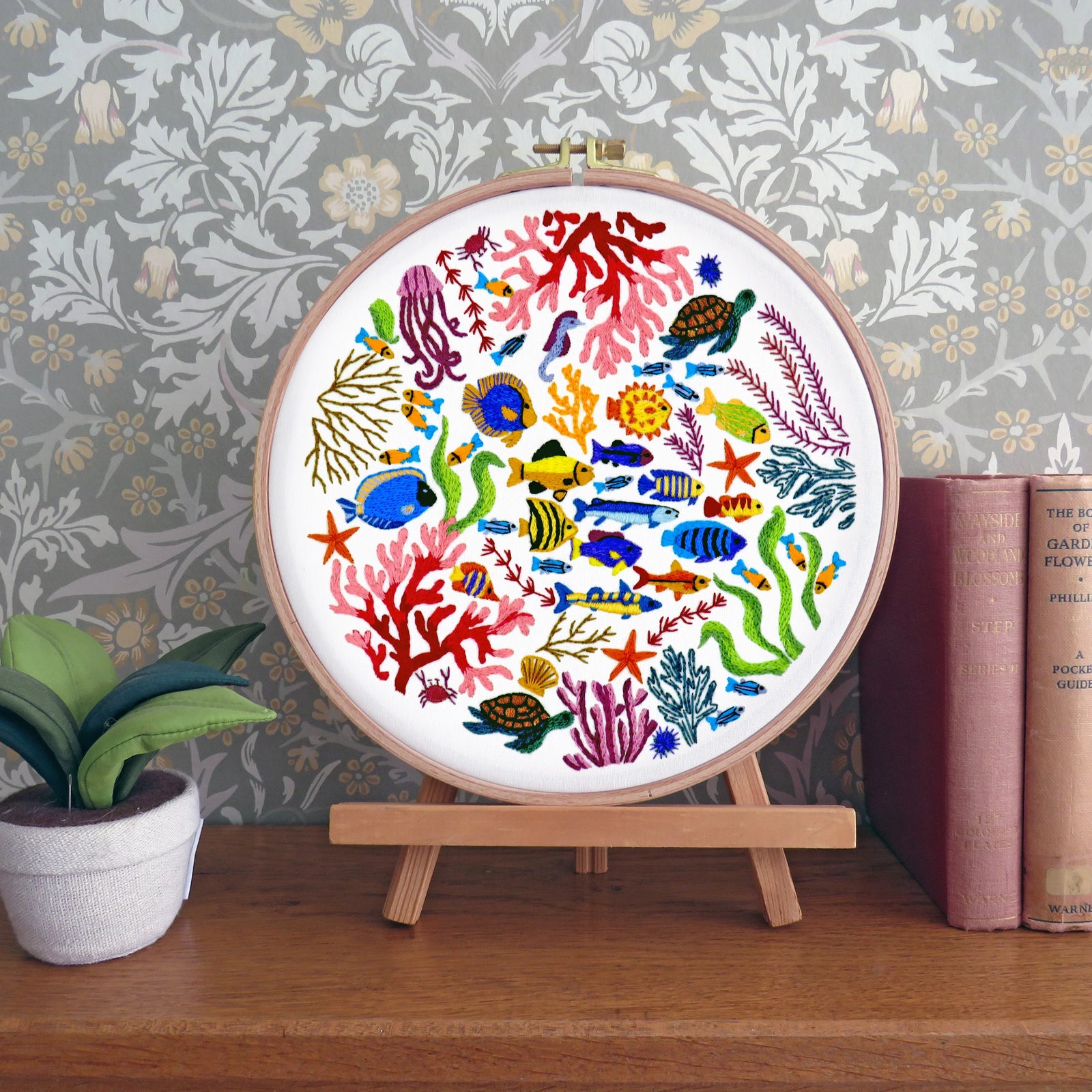 Decorative embroidery hoop with colorful underwater scene on a wooden stand against a floral wallpaper background.