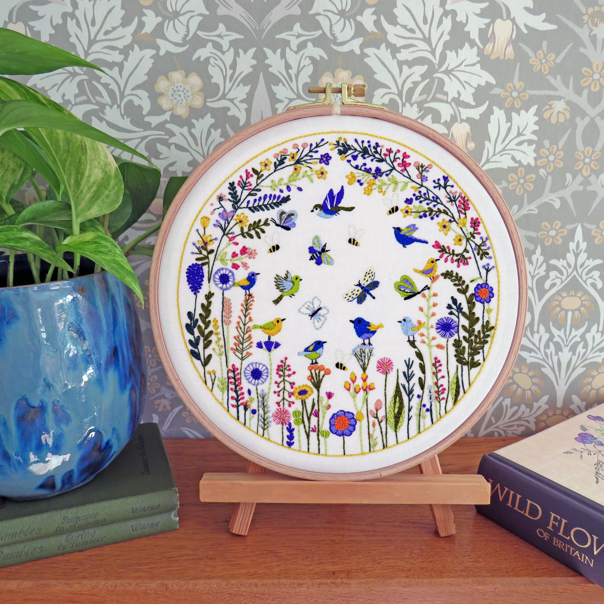 A circular embroidery hoop displaying a colorful floral and bird design, set on a wooden stand on a tabletop with a potted plant and book in the background.