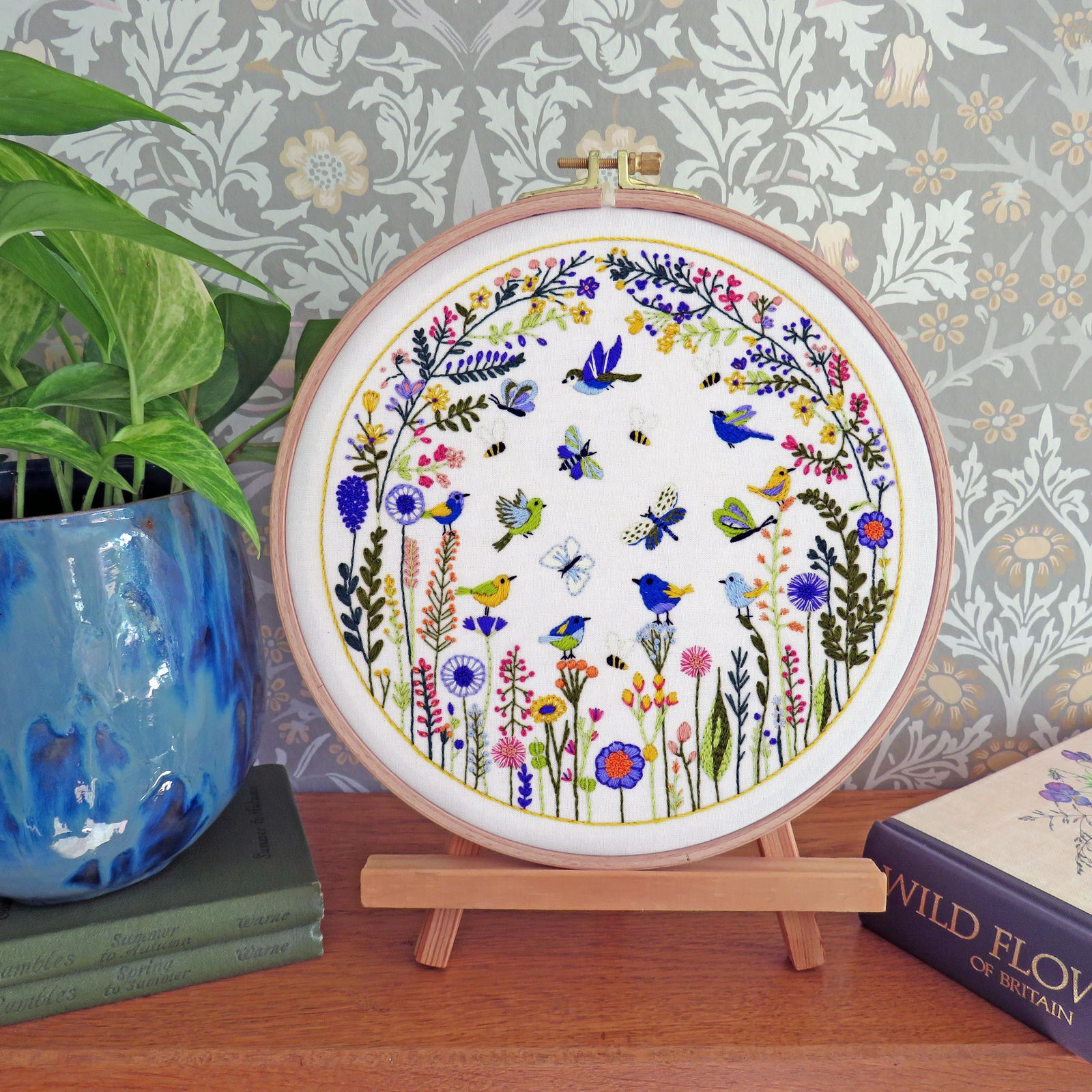 A circular embroidery hoop displaying a colorful floral and bird design, set on a wooden stand on a tabletop with a potted plant and book in the background.