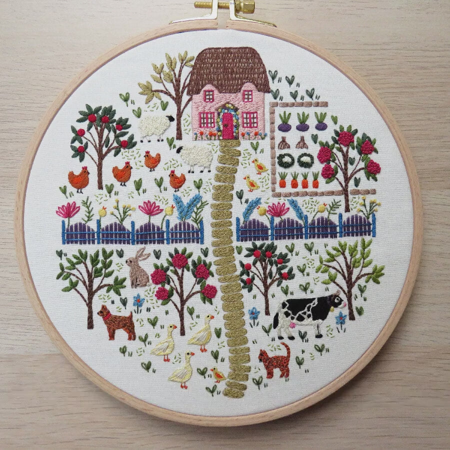 Embroidery hoop with a colorful garden scene including a house, animals, and plants on a purple background.