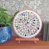 A circular embroidery hoop displaying a colorful floral mandala design on white thread, mounted on a small wooden stand on a wooden shelf. A potted green plant is on the left, and a stack of books is on the right, against a patterned wallpaper background.