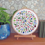 A circular embroidery hoop displaying a colorful floral mandala design on white thread, mounted on a small wooden stand on a wooden shelf. A potted green plant is on the left, and a stack of books is on the right, against a patterned wallpaper background.