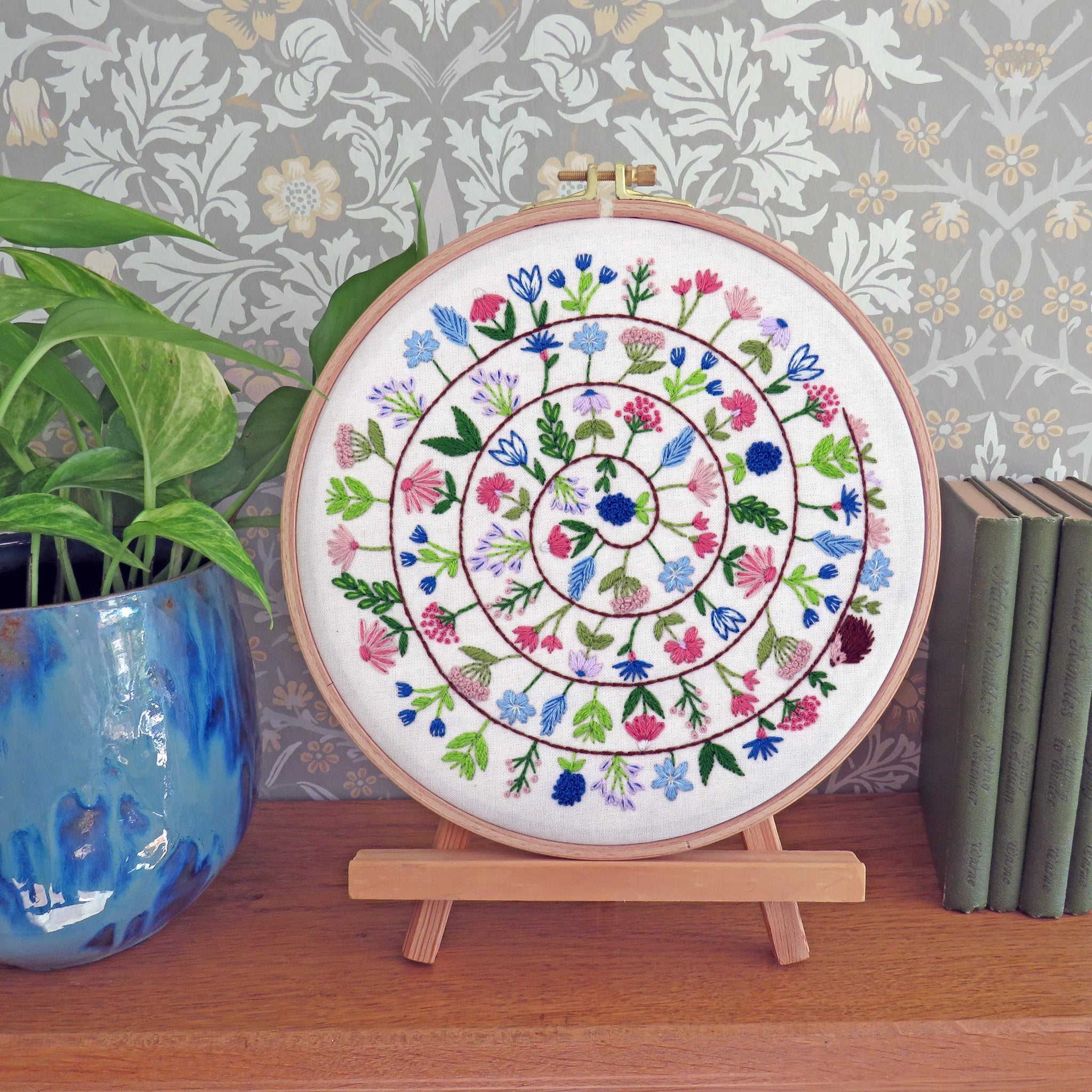 A circular embroidery hoop displaying a colorful floral mandala design on white thread, mounted on a small wooden stand on a wooden shelf. A potted green plant is on the left, and a stack of books is on the right, against a patterned wallpaper background.