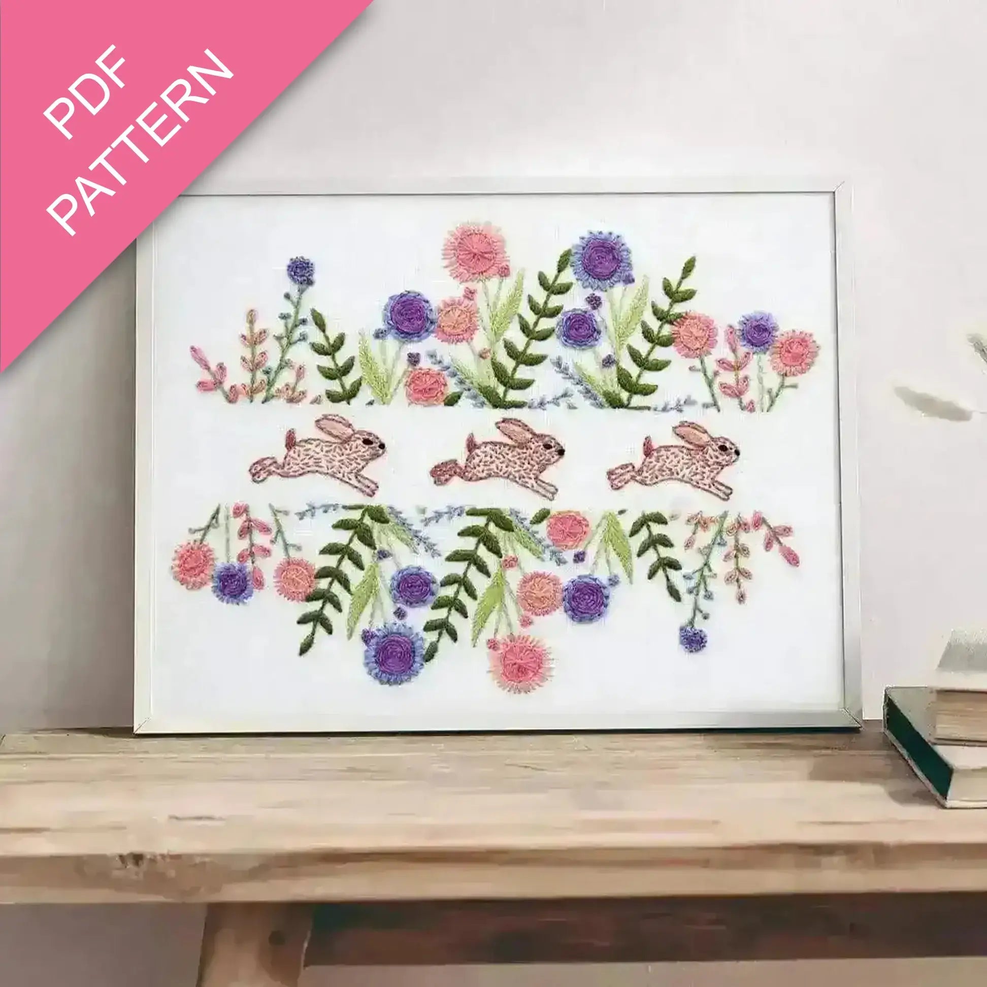 Embroidery design with bunnies and flowers on a white background, framed and displayed on a wooden surface.